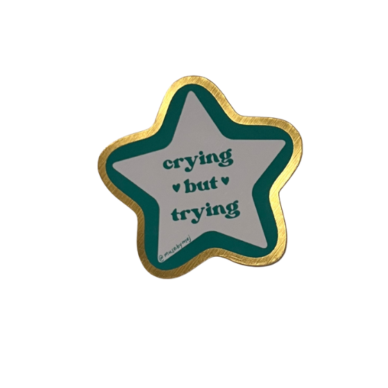 Star-shaped sticker with 'crying but trying' text in aqua written on a white background in a star shape, then the star shape has a border of aqua and another border of shiny gold 