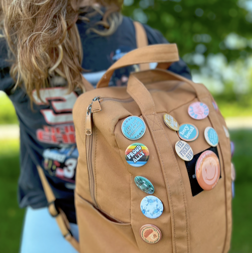 Brown backpack with colorful pins worn by a person outdoors